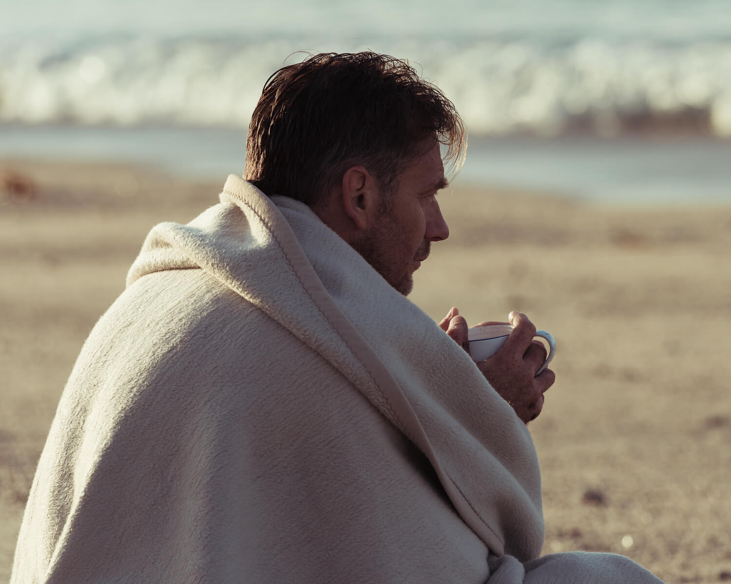 Man sits in blanket on summer beach holding coffee